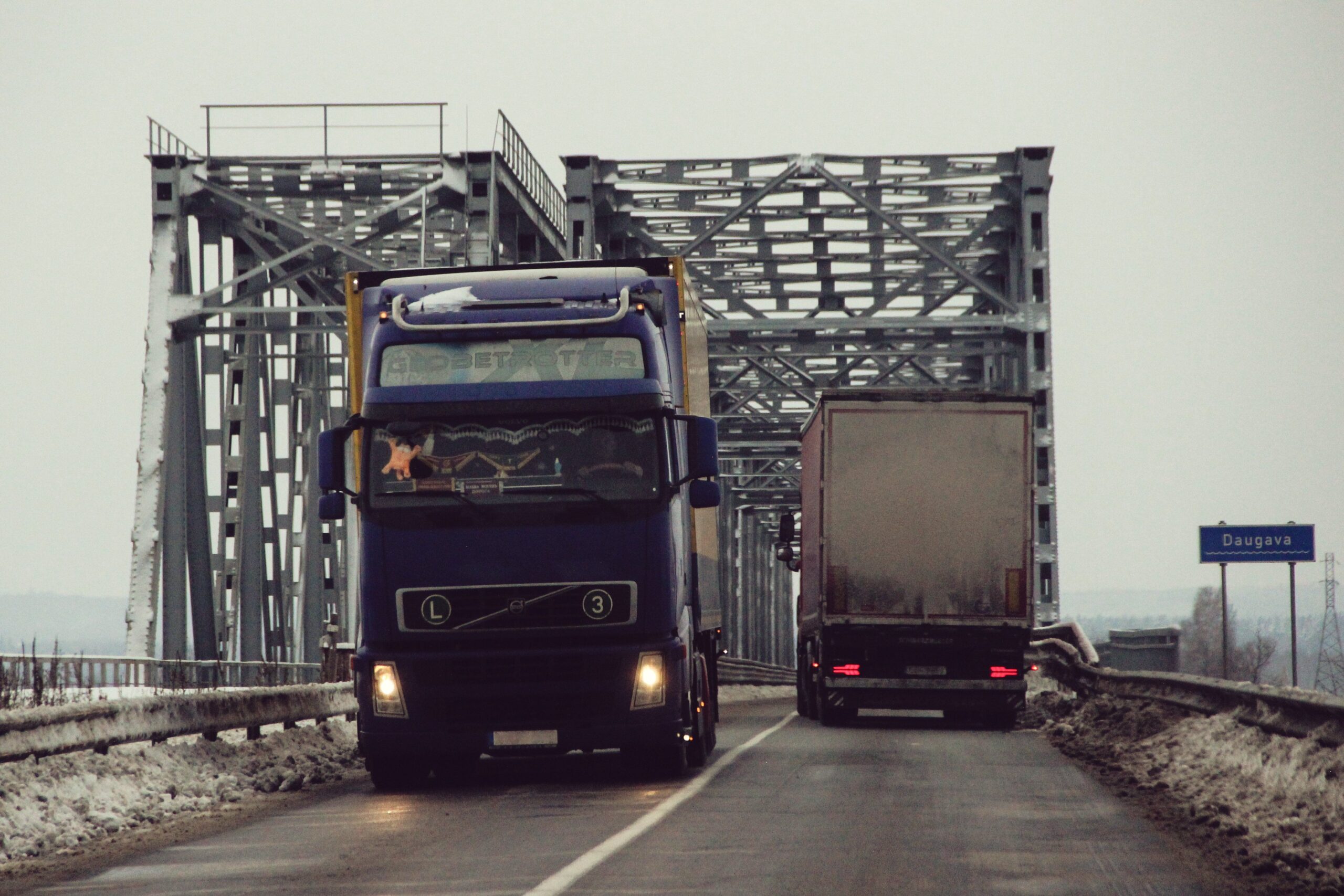 Two trucks navigate a snowy bridge over the Daugava River in Latvia during winter.